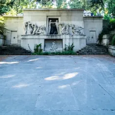 Monumentalna fasada z płaskorzeźbami na cmentarzu Père Lachaise. Kamienne schody prowadzą do wejścia, a wokół rosną drzewa.