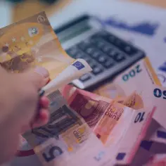 Dłoń trzyma banknot euro, pokazując, jaka waluta we Francji jest używana. W tle kalkulator i wykresy finansowe.