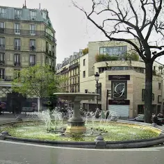 Fontanna na placu Pigalle w Paryżu, otoczona kamienicami i drzewem.