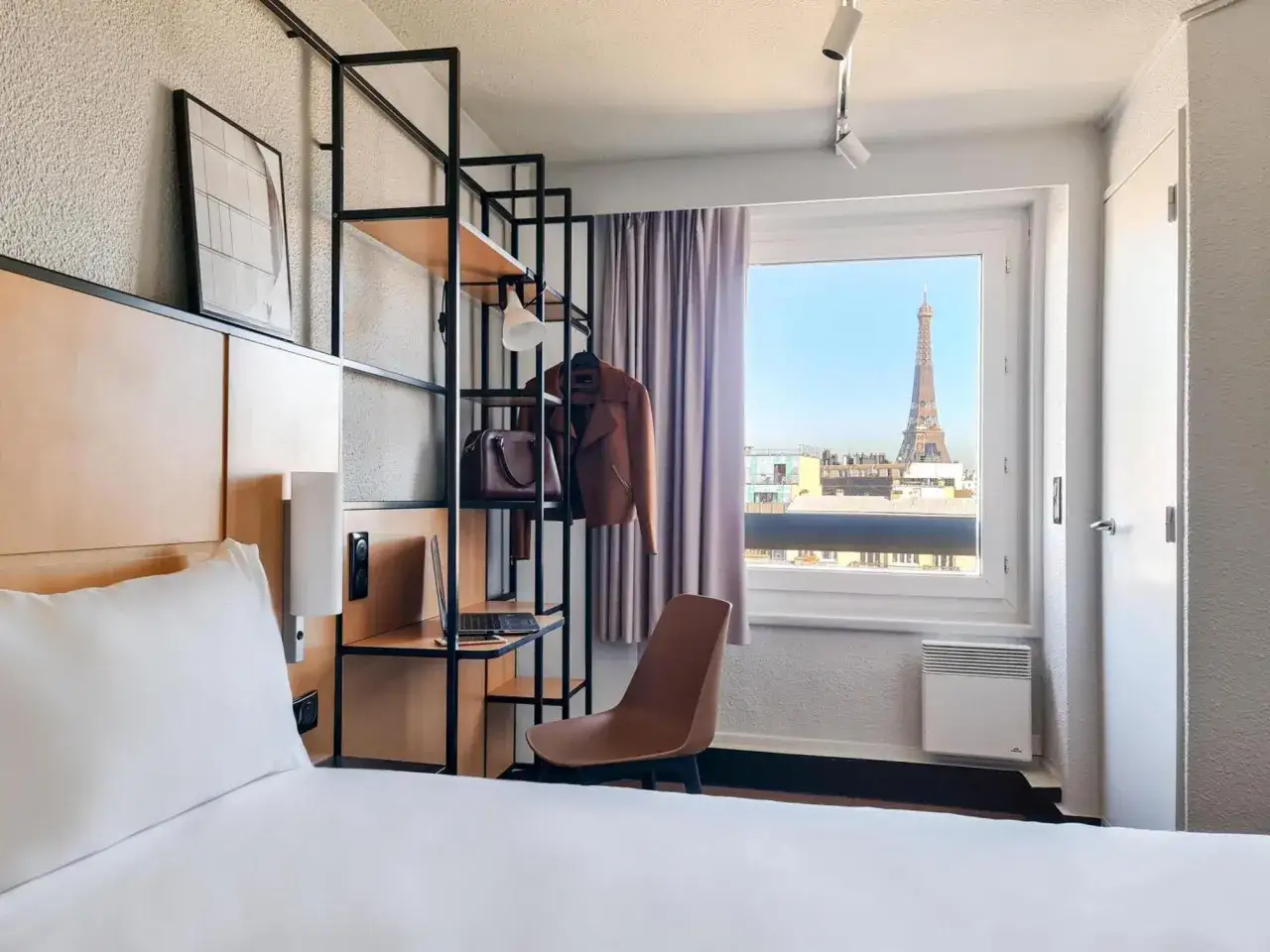 Pokój w Ibis Hotel Paris z widokiem na Wieżę Eiffla. Wygodne łóżko, regał z kurtką i torbą, krzesło.