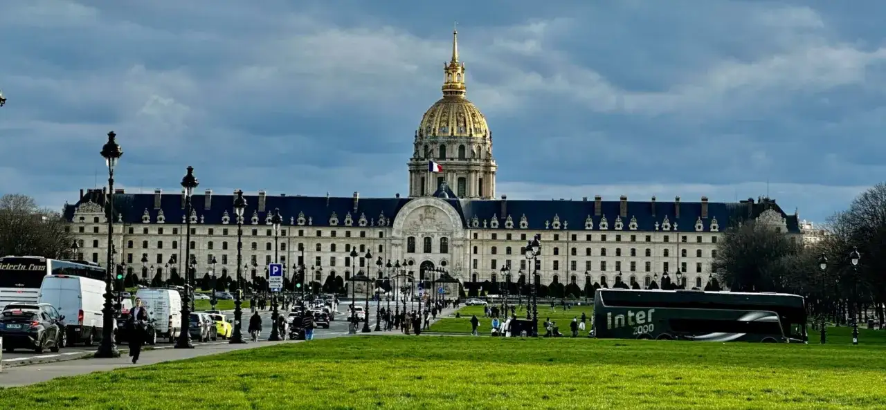 Złota kopuła Les Invalides góruje nad placem w Paryżu. Ruch uliczny i zielony trawnik przed monumentalnym budynkiem.