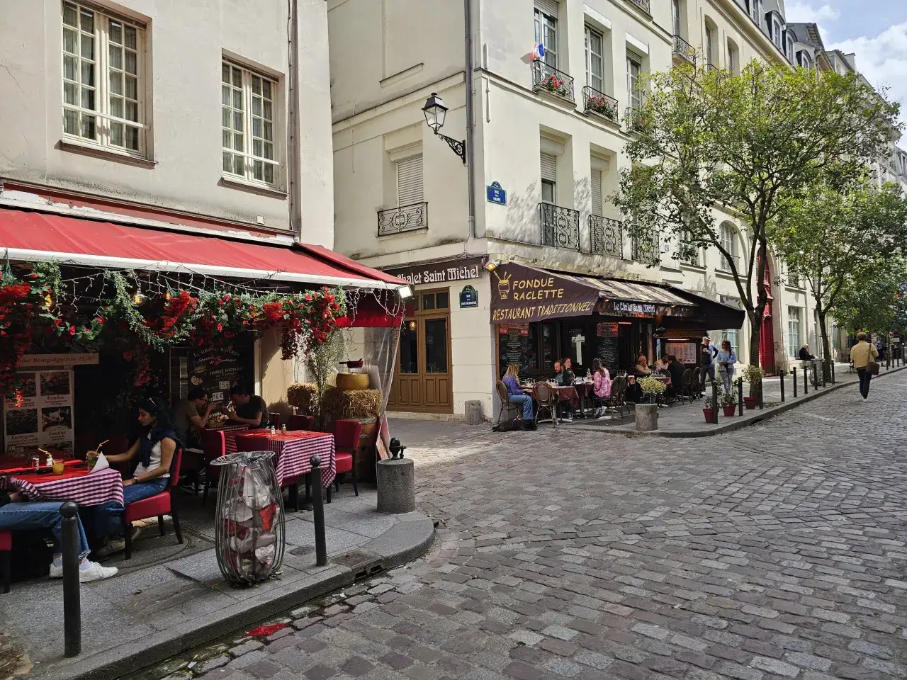 Urokliwa uliczka w paryskiej Dzielnicy Łacińskiej. Stoliki restauracyjne na brukowanym chodniku, ozdobione czerwonymi kwiatami i girlandami.