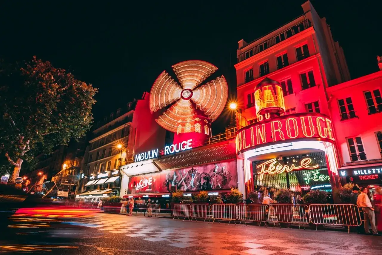 Nocne światła Paryża rozświetlają neonowy szyld kabaretu Moulin Rouge. Wirujący wiatrak na dachu i czerwone budynki tworzą magiczną atmosferę.