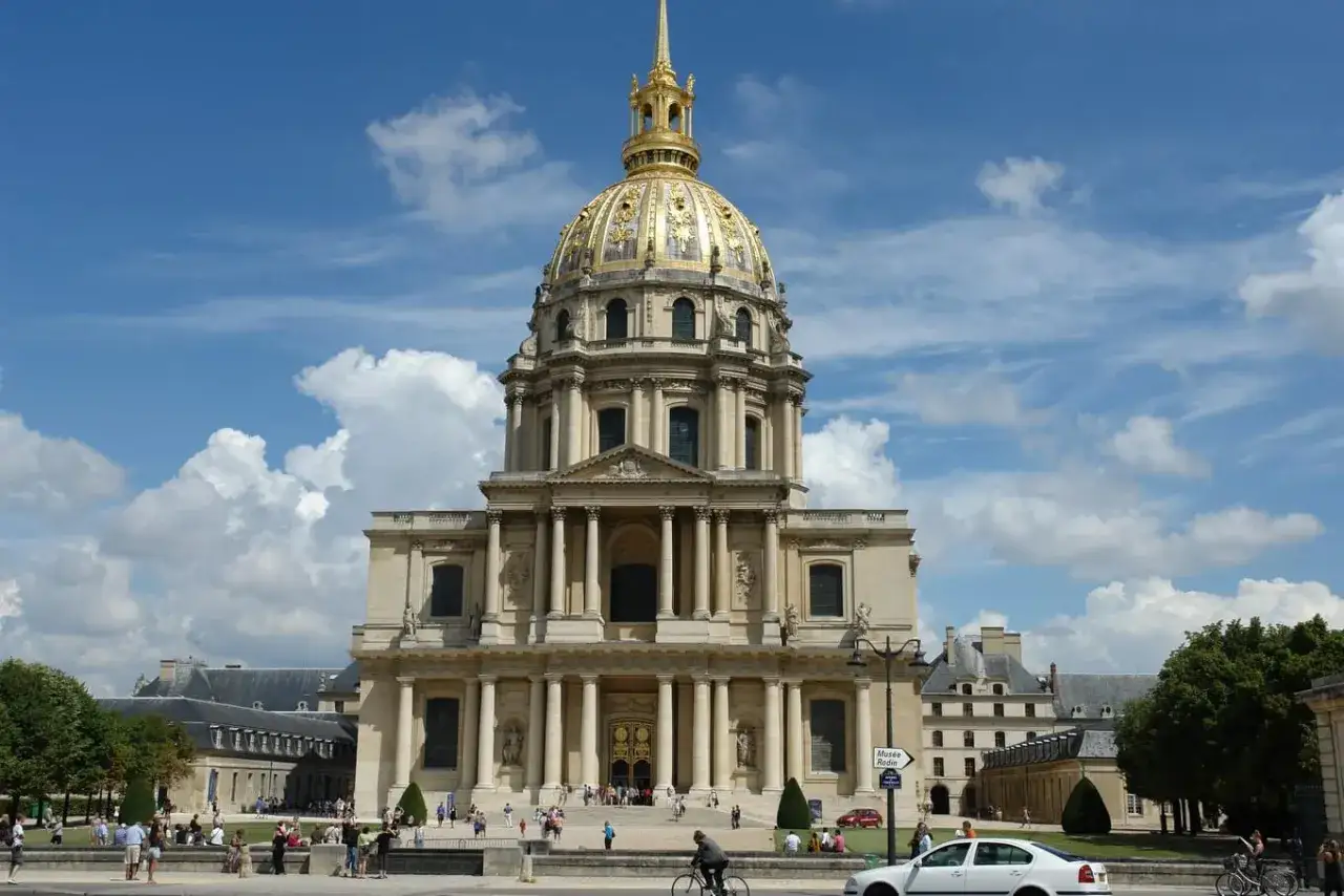 Złota kopuła Les Invalides w Paryżu góruje nad placem, gdzie spacerują ludzie i jeżdżą samochody.